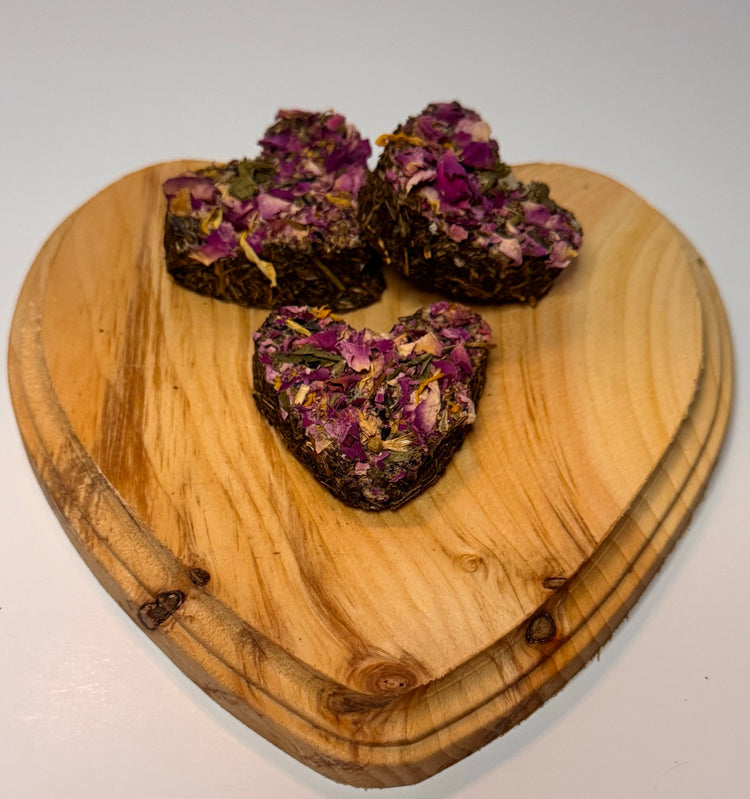 Heart-shaped wooden plate with two heart shaped forage herbal bite treats for rabbits topped with rose petals.