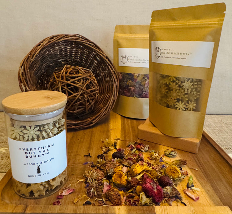 Soft apothecary-style rabbit enrichment display featuring BunBun & Co forage blends, botanical blossom treats in a glass jar, willow enrichment toys, and dried floral forage arranged on a wooden board with warm neutral tones.