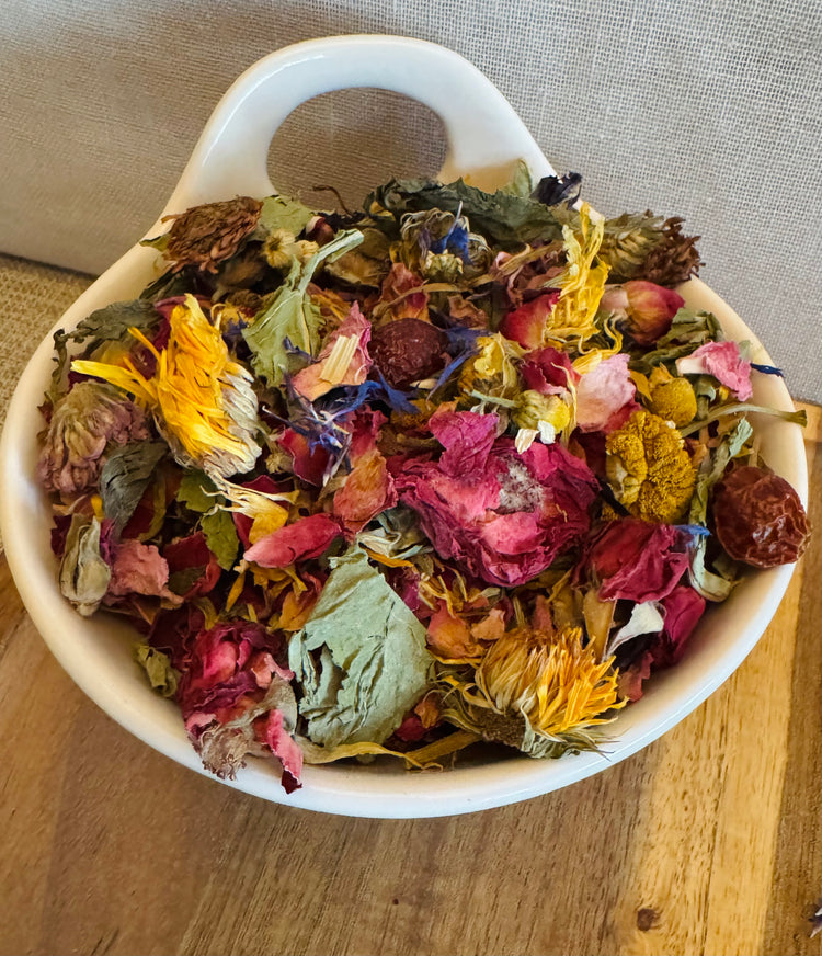 Close-up of colorful dried botanical forage blend for rabbits featuring rose petals, calendula, chamomile, blue cornflower, and leafy herbs styled in a white ceramic bowl on a wooden surface.