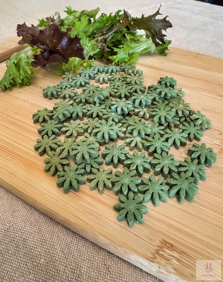 Small green blossom- shaped rabbit treats displayed on a light brown cutting board with fresh greens in the background.