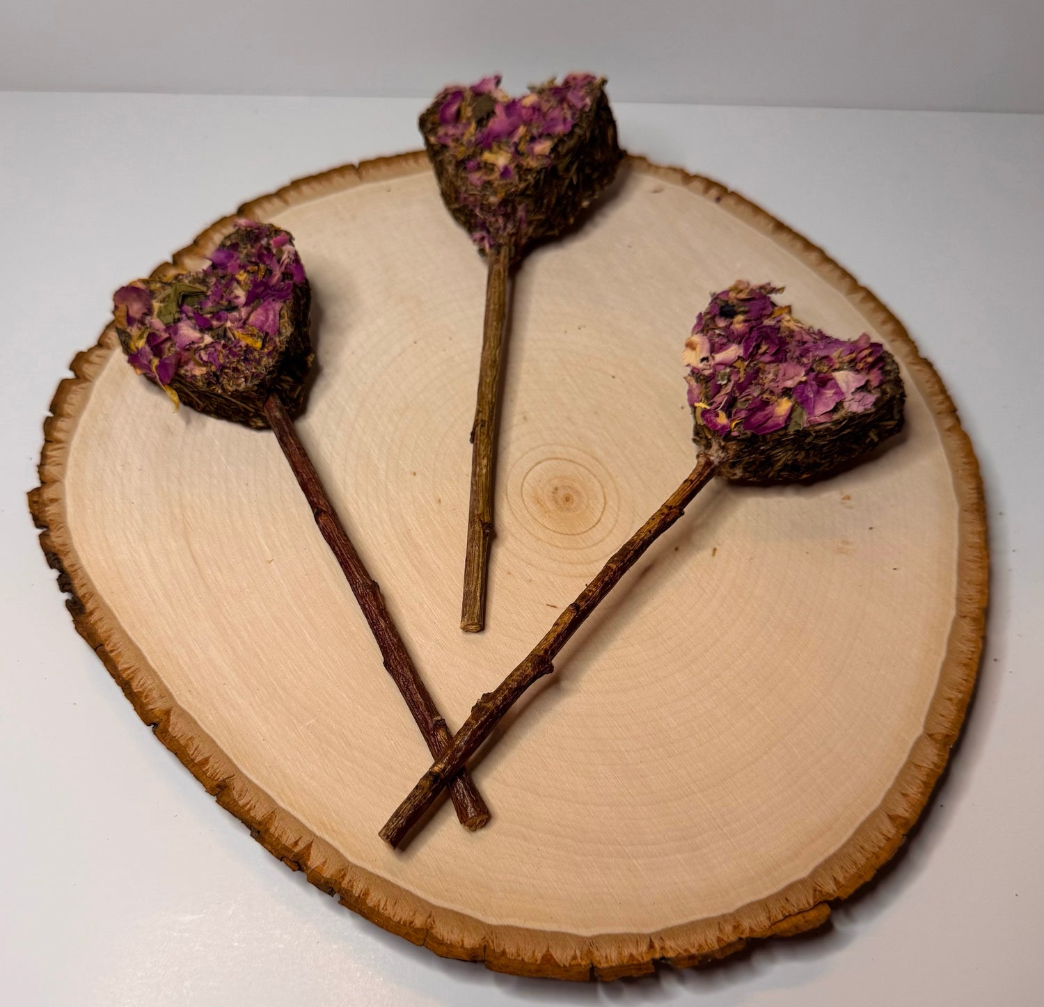 Heart-shaped rabbit forage pops with dried rose petals, botanicals  and a peach wood chew stick handles displayed on natural wood slice