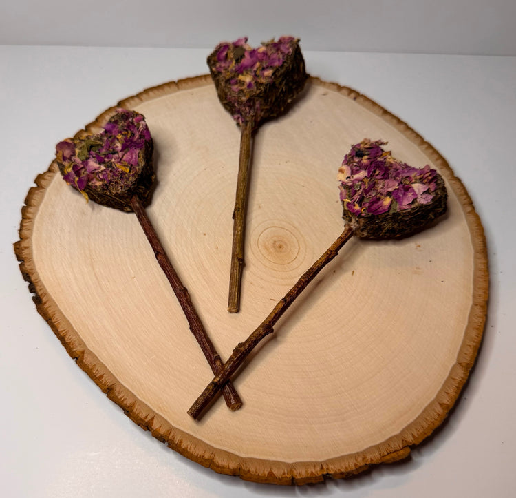 Heart-shaped rabbit forage pops with dried rose petals, botanicals  and a peach wood chew stick handles displayed on natural wood slice