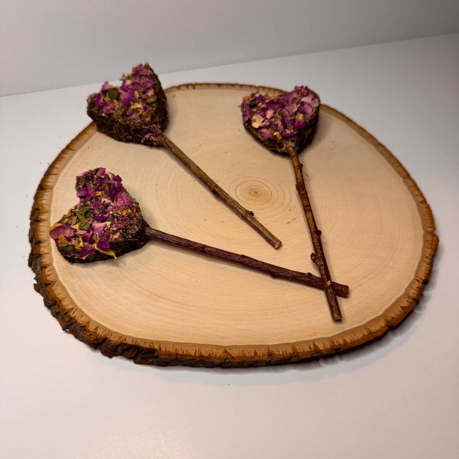 Heart-shaped rabbit forage pops with dried rose petals, botanicals  and a peach wood chew stick handles displayed on natural wood slice