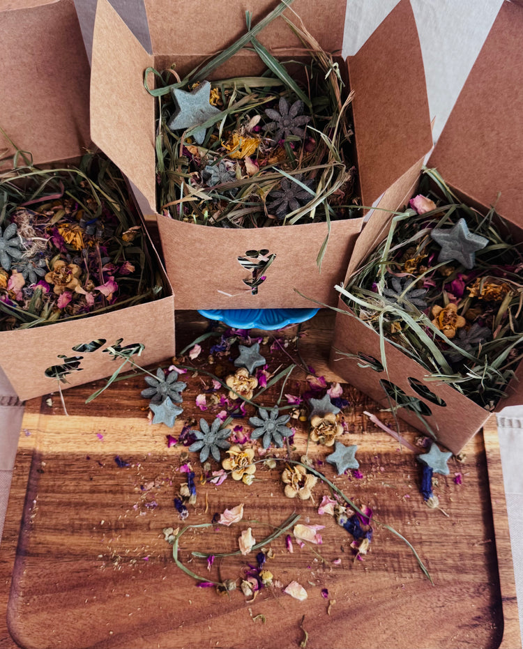 Three open brown kraft paper gift boxes filled with botanicals, herbs and flowers, with scattered star-shaped rabbit treat pieces, arranged on a wooden board with loose dried botanicals scattered in front.