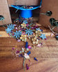 A styled flat-lay of dried botanicals and small flower shaped rabbit treats scattered on a wooden surface, with brown cardboard boxes and a blue pedistool in the background.