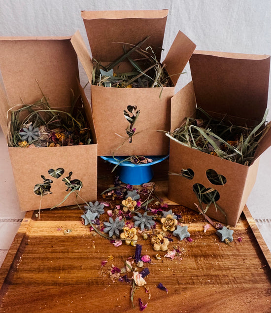 Three brown kraft paper rabbit forage  boxes filled with hay grass and flower petals, arranged on a wooden tray with loose dried botanicals scattered and rabbit treats in front.