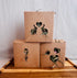 Three small brown kraft paper PEEK A BUN FORAGE boxes stacked in a pyramid arrangement on a wooden tray, each with a decorative cutout showing green hay and botanicals.