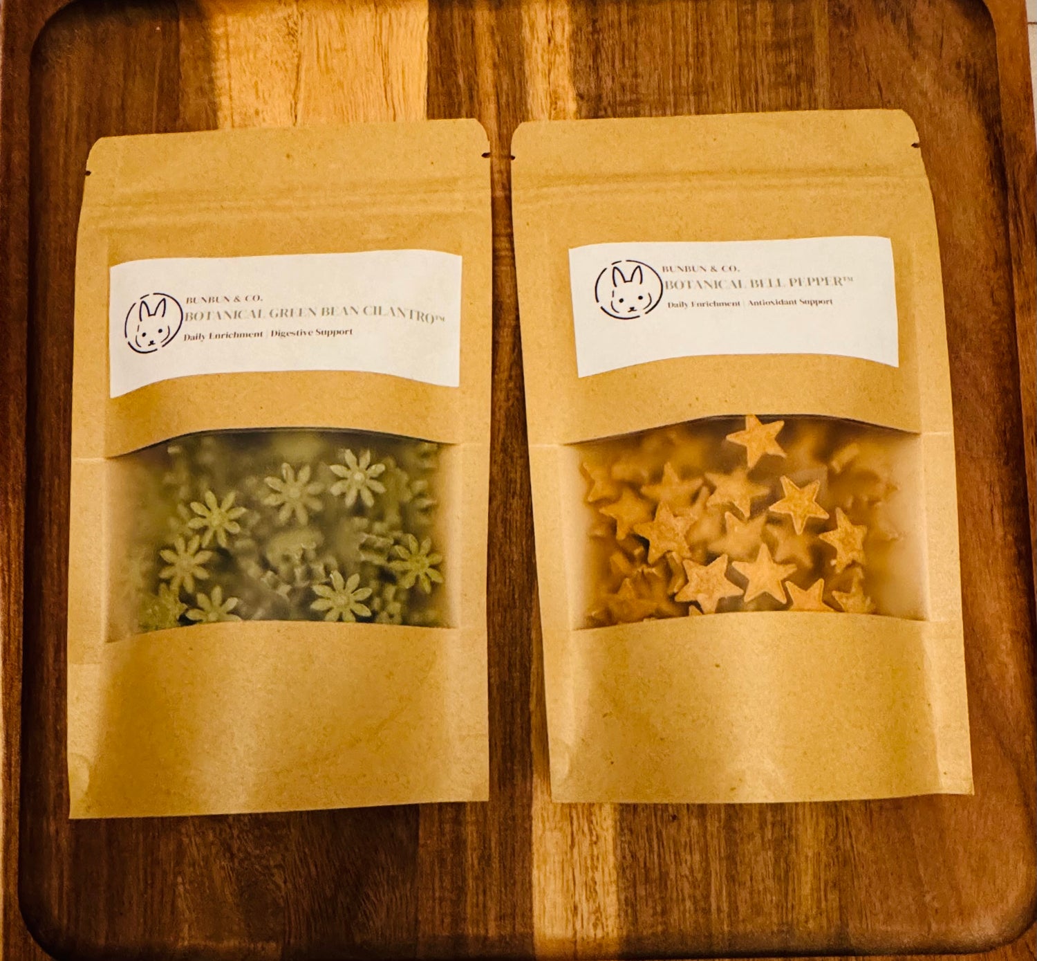 Two  BunBun & Co. brown kraft pouches  with star & blossom shaped rabbit treats on a wooden board.