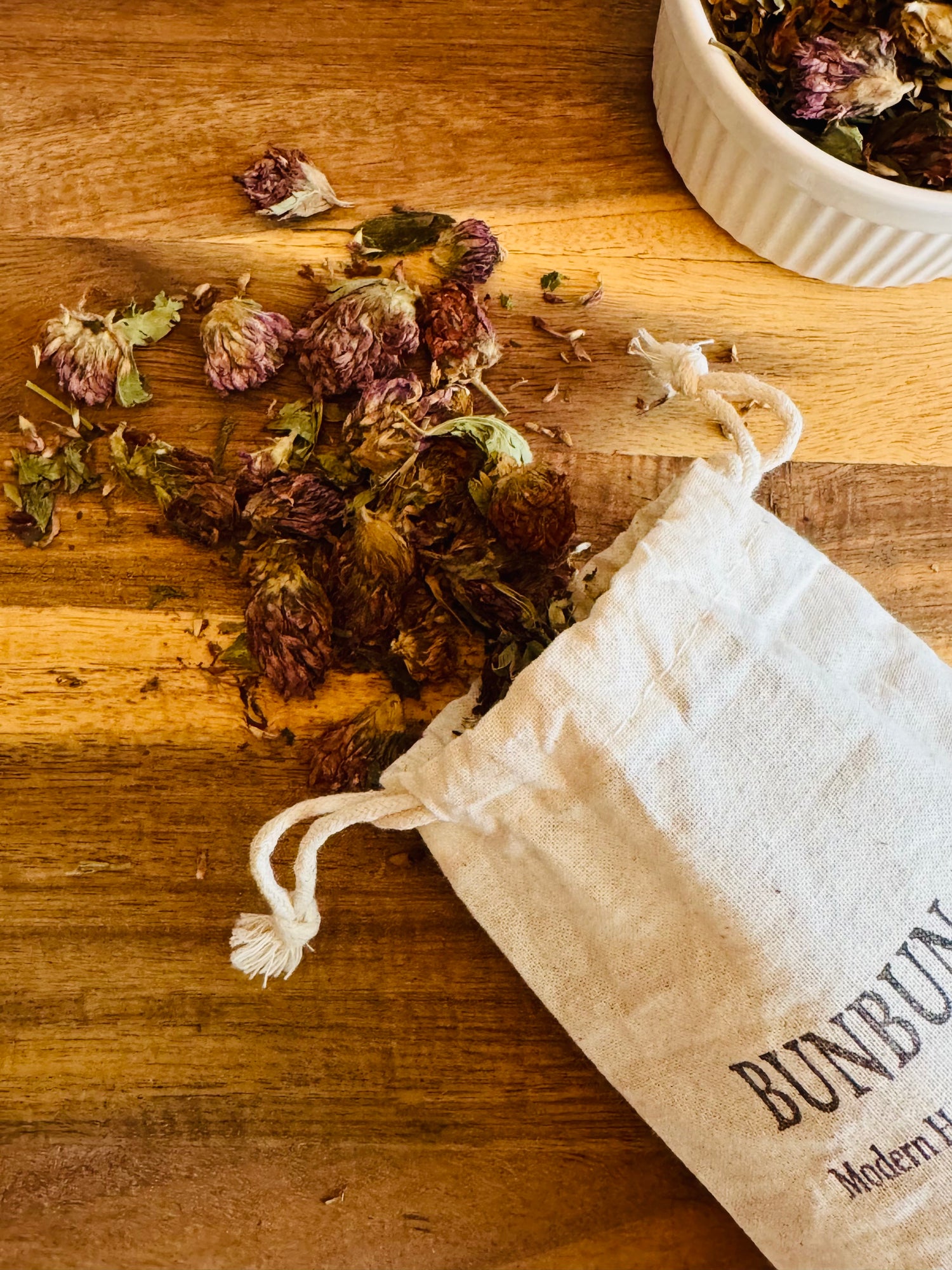 Dried red clover blossoms  spilling out onto a wooden surface with a white cotton &