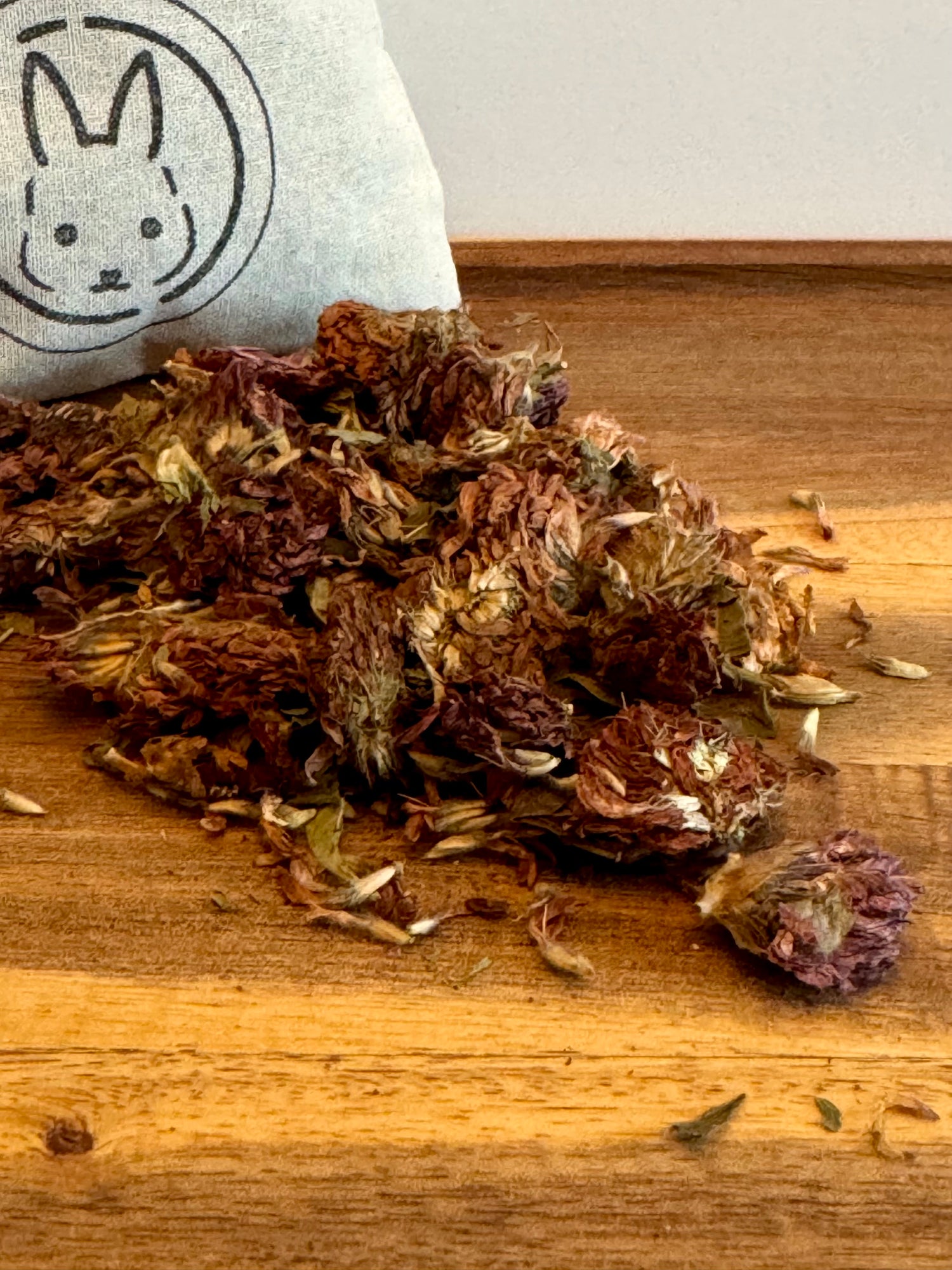 Dried red clover blossoms on a wooden surface with a white cottom bag  featuring a rabbit design in the background.