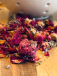 Dried red rose buds and petals on a wooden surface with a white ceramic colander in the background.