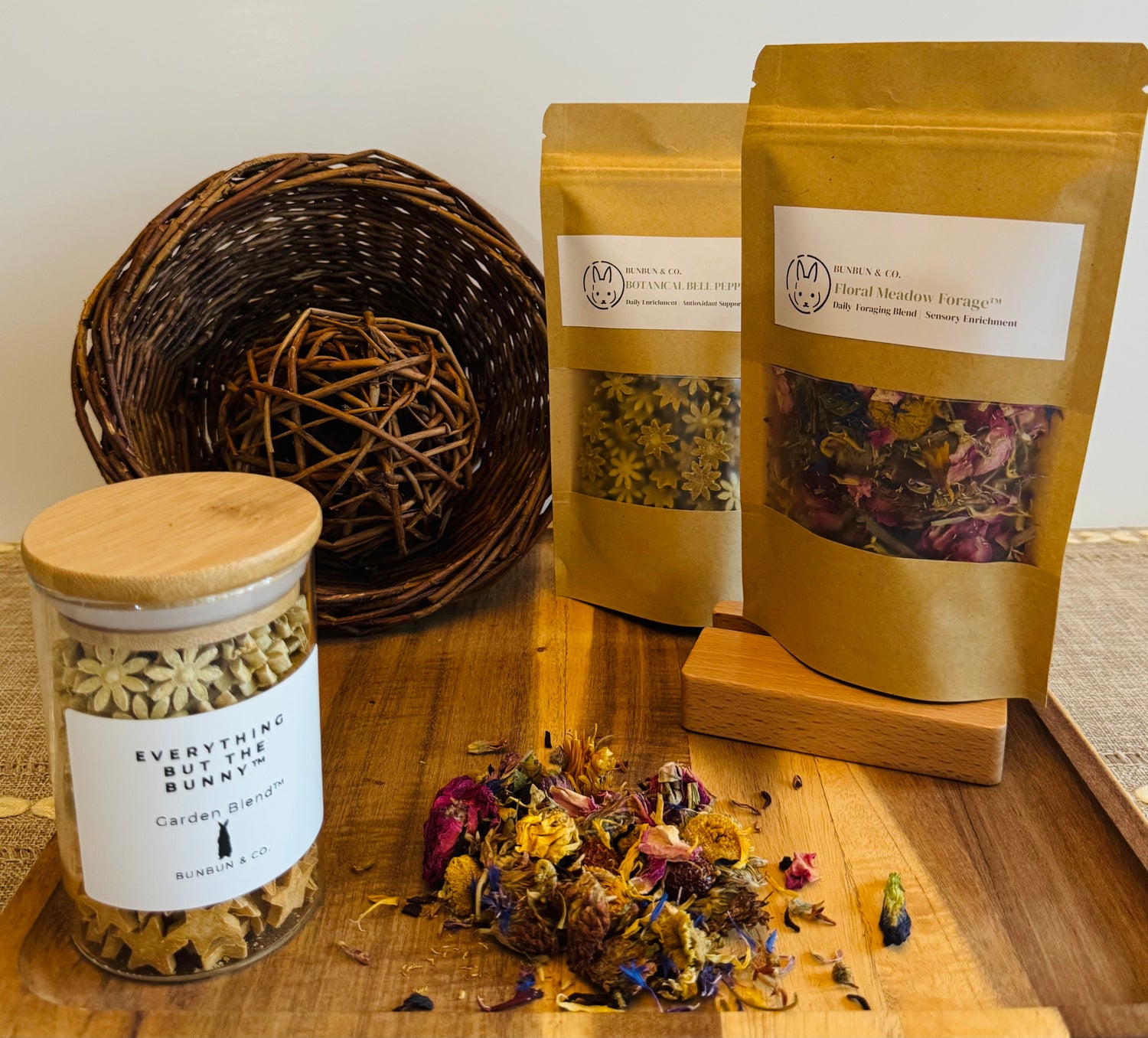 Soft apothecary-style rabbit enrichment display featuring BunBun & Co forage blends, botanical blossom treats in a glass jar, willow enrichment toys, and dried floral forage arranged on a wooden board with warm neutral tones.