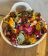 Close-up of colorful dried botanical forage blend for rabbits featuring rose petals, calendula, chamomile, blue cornflower, and leafy herbs styled in a white ceramic bowl on a wooden surface.